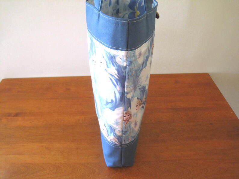 Puede incluir: Un bolso tote alto con la parte superior e inferior azul claro, y un centro estampado en blanco y azul. El estampado parece representar personajes de la pel&iacute;cula Frozen. El bolso est&aacute; sobre una superficie de madera.