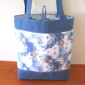 Puede incluir: Un bolso tote azul con un estampado colorido que presenta a una princesa con un vestido azul. El bolso tiene asas y ribetes azules. El forro interior es un patr&oacute;n azul claro y blanco.