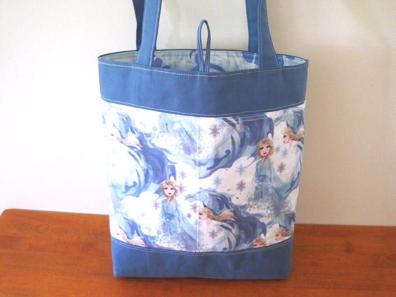 Puede incluir: Un bolso tote azul con un estampado colorido que presenta a una princesa con un vestido azul. El bolso tiene asas y ribetes azules. El forro interior es un patr&oacute;n azul claro y blanco.