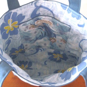 Puede incluir: Un bolso tote azul con forro estampado floral azul y de personajes. El bolso tiene asas azules y un peque&ntilde;o bolsillo interior. El interior presenta un estampado floral azul claro y blanco con detalles amarillos. El bolsillo tiene un estampado de personajes.