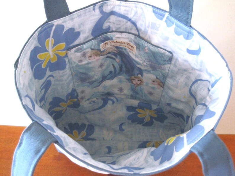 Puede incluir: Un bolso tote azul con forro estampado floral azul y de personajes. El bolso tiene asas azules y un peque&ntilde;o bolsillo interior. El interior presenta un estampado floral azul claro y blanco con detalles amarillos. El bolsillo tiene un estampado de personajes.