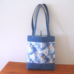 Puede incluir: Un bolso tote azul con forma rectangular y dos asas. El bolso presenta una tela estampada con im&aacute;genes de una princesa en tonos azules y blancos. La parte superior e inferior del bolso son de color azul liso.