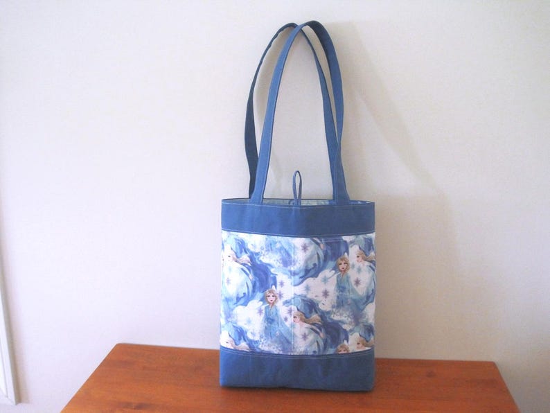 Puede incluir: Un bolso tote azul con forma rectangular y dos asas. El bolso presenta una tela estampada con im&aacute;genes de una princesa en tonos azules y blancos. La parte superior e inferior del bolso son de color azul liso.