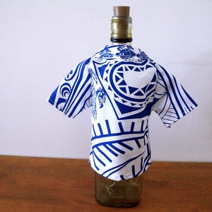 May include: A glass bottle wearing a short-sleeved shirt cover featuring a white and blue tribal-inspired pattern. The shirt has a collar and covers the upper portion of the bottle, which is topped with a brown cork stopper.