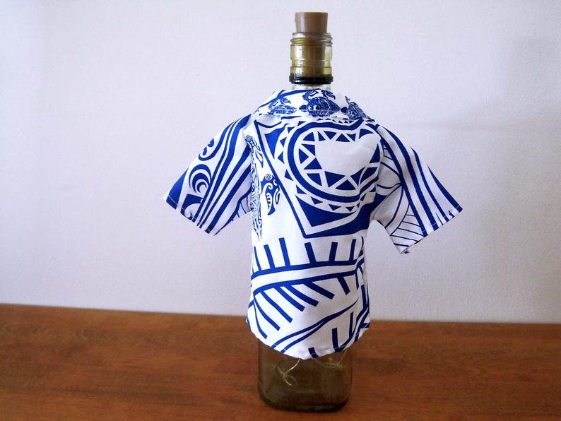 May include: A glass bottle wearing a short-sleeved shirt cover featuring a white and blue tribal-inspired pattern. The shirt has a collar and covers the upper portion of the bottle, which is topped with a brown cork stopper.