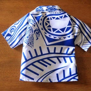 May include: A short-sleeved button-down shirt featuring a white base with a bold blue Polynesian-inspired tribal print. The pattern includes stylized sea turtles and geometric shapes across the collar, sleeves, and body of the garment.