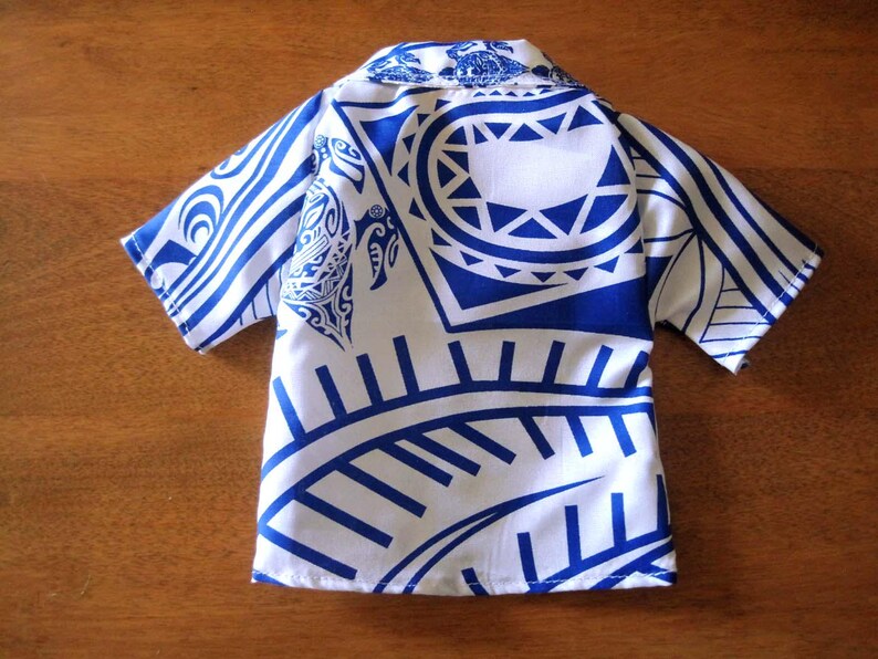 May include: A short-sleeved button-down shirt featuring a white base with a bold blue Polynesian-inspired tribal print. The pattern includes stylized sea turtles and geometric shapes across the collar, sleeves, and body of the garment.