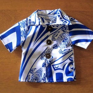 May include: A short-sleeved button-down shirt featuring a vibrant blue and white tribal-inspired pattern with sea turtle motifs. The garment includes a classic collar and three dark brown circular buttons down the center front placket.