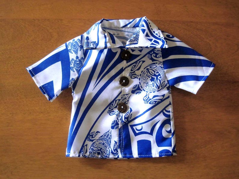 May include: A short-sleeved button-down shirt featuring a vibrant blue and white tribal-inspired pattern with sea turtle motifs. The garment includes a classic collar and three dark brown circular buttons down the center front placket.