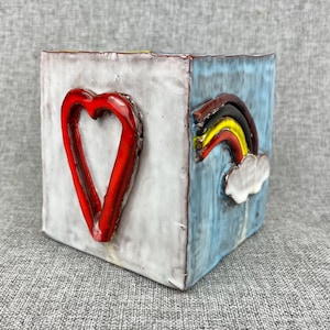 May include: A ceramic planter with a white and blue glaze. One side features a red heart and the other side features a rainbow with a white cloud.