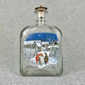May include: A clear glass bottle with a cork stopper and a winter scene painted on the front. The scene depicts a gnome in a red hat and a cat walking through a snowy village.