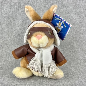 May include: A plush bunny wearing a brown aviator jacket, goggles, and a white scarf. The bunny has long ears and a pink nose. A tag reads "Grumpy Bunny" with a cartoon bunny illustration.