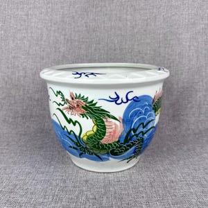 May include: White ceramic planter with a hand-painted dragon design. The dragon is green, pink, and yellow, set against blue waves and accents. The planter has a decorative rim and is suitable for indoor or outdoor use.
