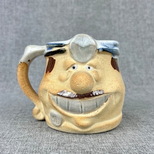 May include: A ceramic mug shaped like a cartoon doctor with a blue and white hat, brown eyebrows, a large brown mustache, and a wide, toothy smile.