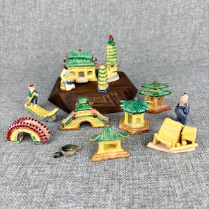 May include: A collection of miniature ceramic Asian-style buildings and structures. The set includes pagodas, bridges, and figures, all painted in vibrant colors like green, yellow, red, and blue. The pieces are arranged on a gray surface.