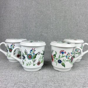 May include: Four white ceramic mugs with lids, each featuring a floral pattern in shades of pink, blue, green, and yellow. The mugs have a simple, elegant design.