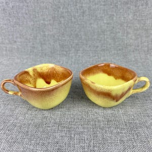 May include: Two yellow ceramic teacups with brown glaze. The cups have a square shape and a handle.