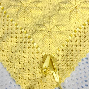 May include: A yellow crocheted baby blanket with a satin ribbon tied in a bow. The blanket has a textured pattern and a delicate crocheted border.