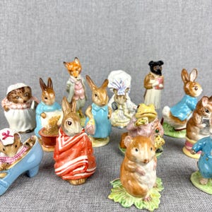 May include: A collection of ceramic figurines featuring various animal characters. The figurines include rabbits, a fox, a frog, and a mouse, all dressed in detailed clothing. The color palette includes shades of blue, red, white, and brown.