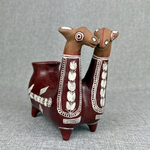May include: A double-necked terracotta vessel shaped like two stylized animals with long necks. The piece features a deep reddish-brown base color with intricate white painted floral patterns and geometric borders along the necks and body.