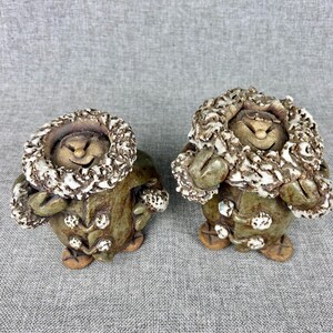 May include: Two ceramic figurines of smiling figures wearing coats with fur-lined hoods. The figures are in a seated position with their hands resting on their laps. The coats are olive green with white accents, and the hoods are a mix of brown and white.
