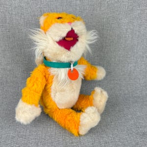 May include: A plush orange and cream-colored cat toy with a green collar and orange tag. The cat has a wide open mouth with a pink interior and a small orange tongue. The toy is sitting on a gray surface.