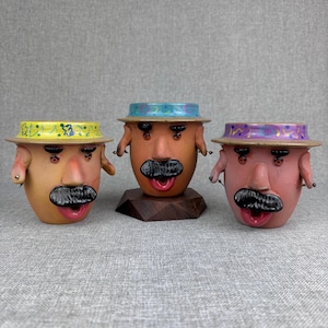 May include: Three ceramic mugs shaped like faces with mustaches, ears, and noses. Each mug wears a wide-brimmed hat with a colorful pattern. The left mug is tan, the center mug is brown on a wooden base, and the right mug is pinkish-tan.