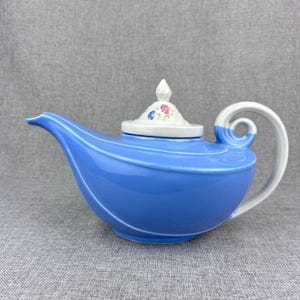 May include: A blue ceramic teapot shaped like a genie lamp with a white lid and floral design. The teapot has a curved spout and a handle that curls around the top.