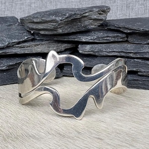 May include: A polished silver cuff bracelet featuring an open, fluid, and abstract wave-like design. The metal surface is smooth and reflective, showcasing a continuous, interconnected pattern that wraps around the wrist with organic, curved edges.