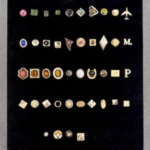 May include: A collection of assorted lapel pins and tie tacks arranged in rows on a black velvet display board. The pins feature various shapes, including geometric, circular, and letter designs, with metallic finishes, gemstones, and enamel accents.