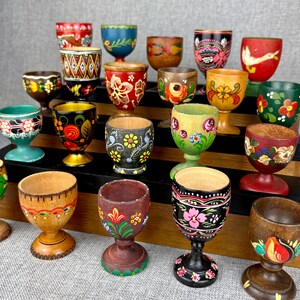 May include: A collection of hand-painted wooden egg cups in various colors and designs. The egg cups feature floral patterns, geometric shapes, and other decorative elements. The collection is displayed on a tiered wooden shelf.