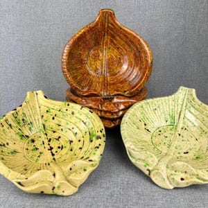 May include: A set of ceramic leaf-shaped dishes in shades of brown, yellow, and green. The dishes have a textured surface with speckled detailing. The stack of dishes is in the background, with individual dishes in the foreground.