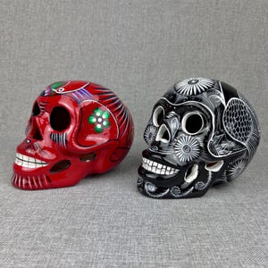 May include: Two decorative skull figurines, one in glossy red with green and white floral accents, and one in glossy black with intricate white line patterns. Both skulls feature hollow eye sockets, nasal cavities, and detailed teeth carvings.