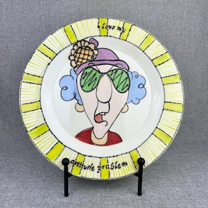 May include: A white ceramic plate with a yellow and white striped border. The center of the plate features a cartoon illustration of a woman with blue hair, green sunglasses, and a purple hat. The text "I love my attitude problem" is written on the plate.