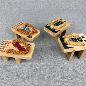 May include: Four miniature wooden geta sandals with rope straps. The geta have a rectangular base with two wooden supports. The tops of the geta are decorated with Japanese text and illustrations. The rope straps are a light brown color.
