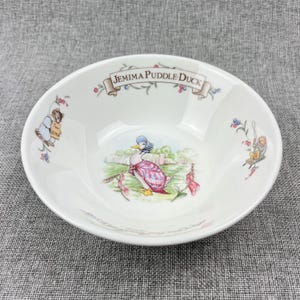 May include: A white ceramic bowl featuring a central illustration of Jemima Puddle-Duck wearing a blue bonnet and pink shawl. The rim displays the text Jemima Puddle-Duck with floral accents, while smaller illustrations of Tom Kitten and other characters appear on the sides.