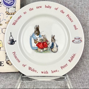 May include: A white ceramic commemorative plate featuring a central illustration of Peter Rabbit and other rabbits. The rim text reads: Welcome to the new baby of the Prince and Princess of Wales, with love, Peter Rabbit. Includes a small tea cup icon.