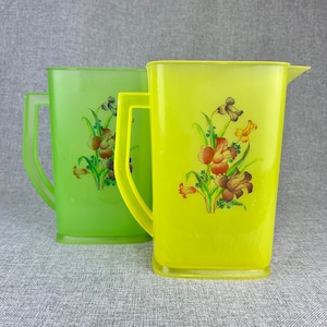 May include: Two plastic pitchers with floral designs. The pitcher on the left is green with a floral design in shades of green, brown, and white. The pitcher on the right is yellow with a floral design in shades of yellow, brown, and green.