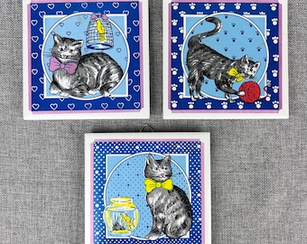 May include: Three square ceramic tiles with cat illustrations. Each tile features a different cat design: one with a pink bow tie and birdcage, another playing with yarn, and the last with a yellow bow tie and fishbowl. The tiles have a blue and white border.