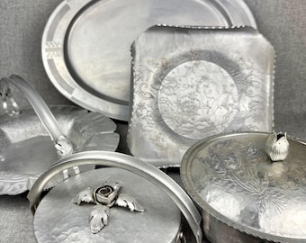 May include: A collection of silver-toned metal serving pieces including an oval platter, a square tray with an embossed floral center, a handled basket, and two round lidded containers featuring decorative metal rose and bud finials.