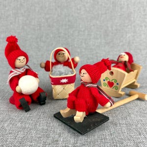 May include: A collection of four wooden Christmas figurines. Each figure is dressed in a red knitted outfit with a matching hat and scarf. One figure holds a white ball, another a basket, and another is in a wooden sled.