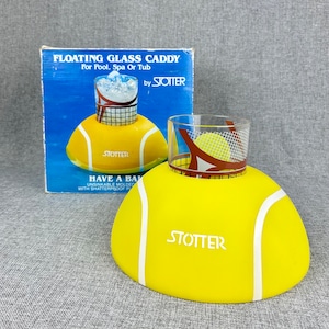 May include: A yellow floating glass caddy shaped like a tennis ball with the word "STOTTER" printed on it. The caddy is designed to hold a glass of beverage and float in a pool or spa. The box shows a glass of ice water in the caddy.