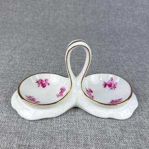 May include: White porcelain salt and pepper shaker set with a pink floral design and gold trim. The shakers are connected by a handle.