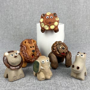 May include: A collection of ceramic animal figurines, including lions, an elephant, and a hippo. The lions have detailed manes, and the elephant has a green ear. The figurines are in various shades of brown and tan, with painted details.