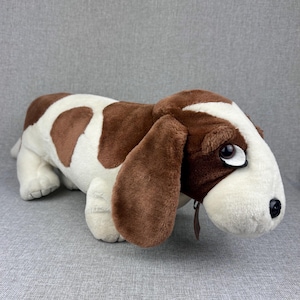 May include: A plush toy shaped like a basset hound dog with a long body, short legs, and large, floppy ears. The stuffed animal features a soft, white and brown spotted coat, a dark brown nose, and large, expressive eyes with white accents.