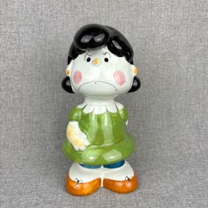 May include: A ceramic figurine of a cartoon character with black hair, a green dress, and orange shoes. The figurine has a sad expression with pink cheeks and a white face. The figure is standing upright.