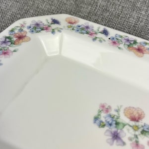 May include: A white rectangular serving dish with a floral design. The dish features a delicate border of pastel-colored flowers, including pink, purple, and yellow blooms. The flowers are arranged in a repeating pattern along the edges of the dish.
