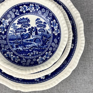 May include: A stack of white and blue ceramic plates. The plates feature a blue and white landscape design in the center, surrounded by floral and geometric patterns. The edges have a scalloped design.