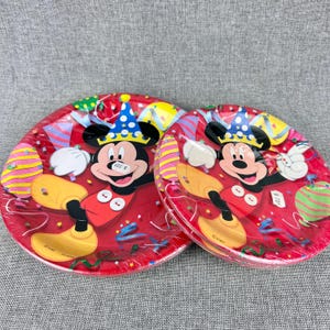 May include: Two red paper plates featuring Mickey Mouse in a party hat, surrounded by balloons and confetti. The plates are round and have a festive design, perfect for a birthday celebration or themed party.