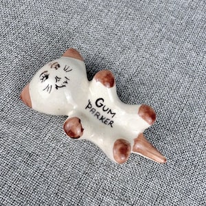 May include: A cream-colored ceramic cat figurine with brown-tipped ears, paws, and tail. The cat has a hand-painted face with black eyes, whiskers, and a nose. The text GUM PARKER is written in black ink on the cat's belly area in a vertical orientation.